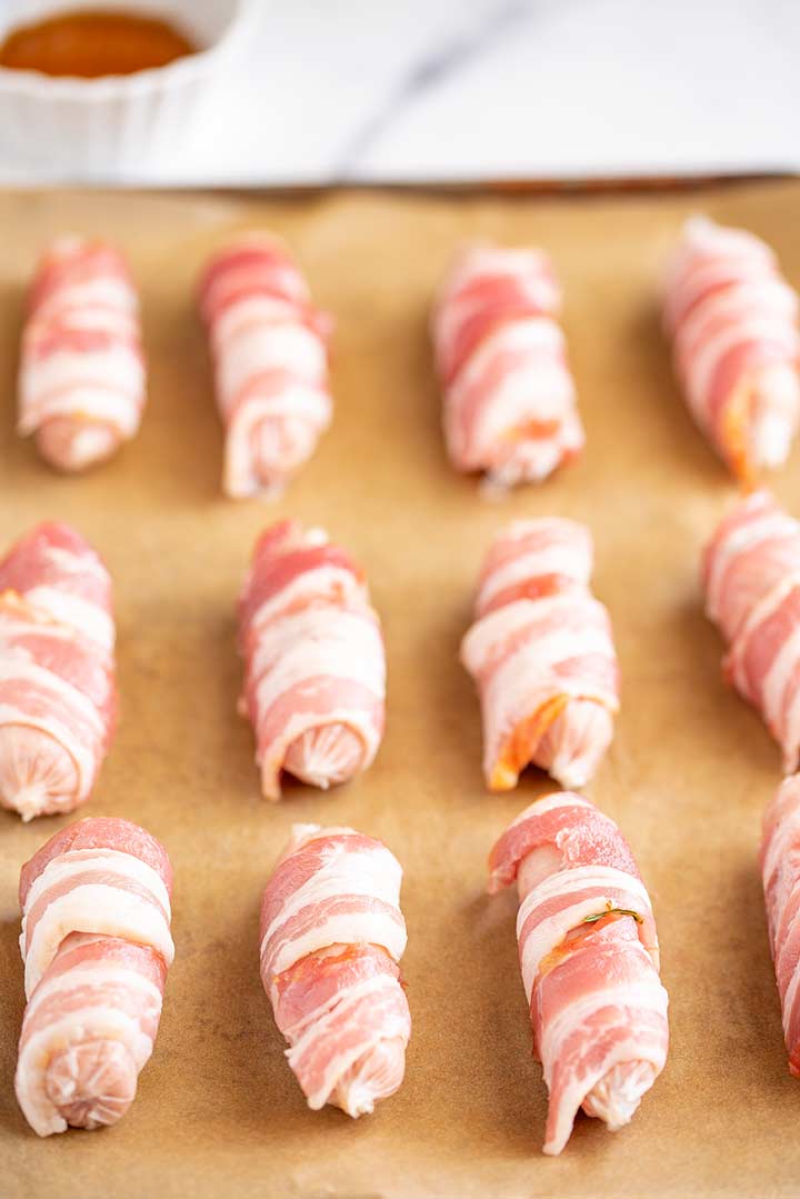 12 raw sausages wrapped in bacon on a lined sheet pan