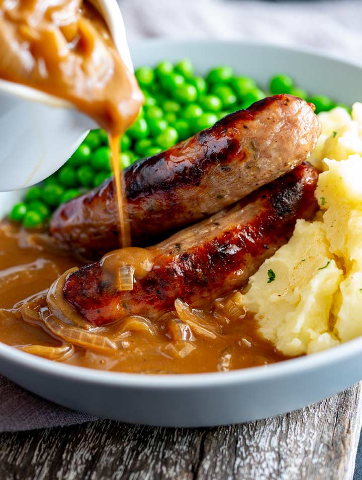 onion gravy being poured over sausages and mashed potato