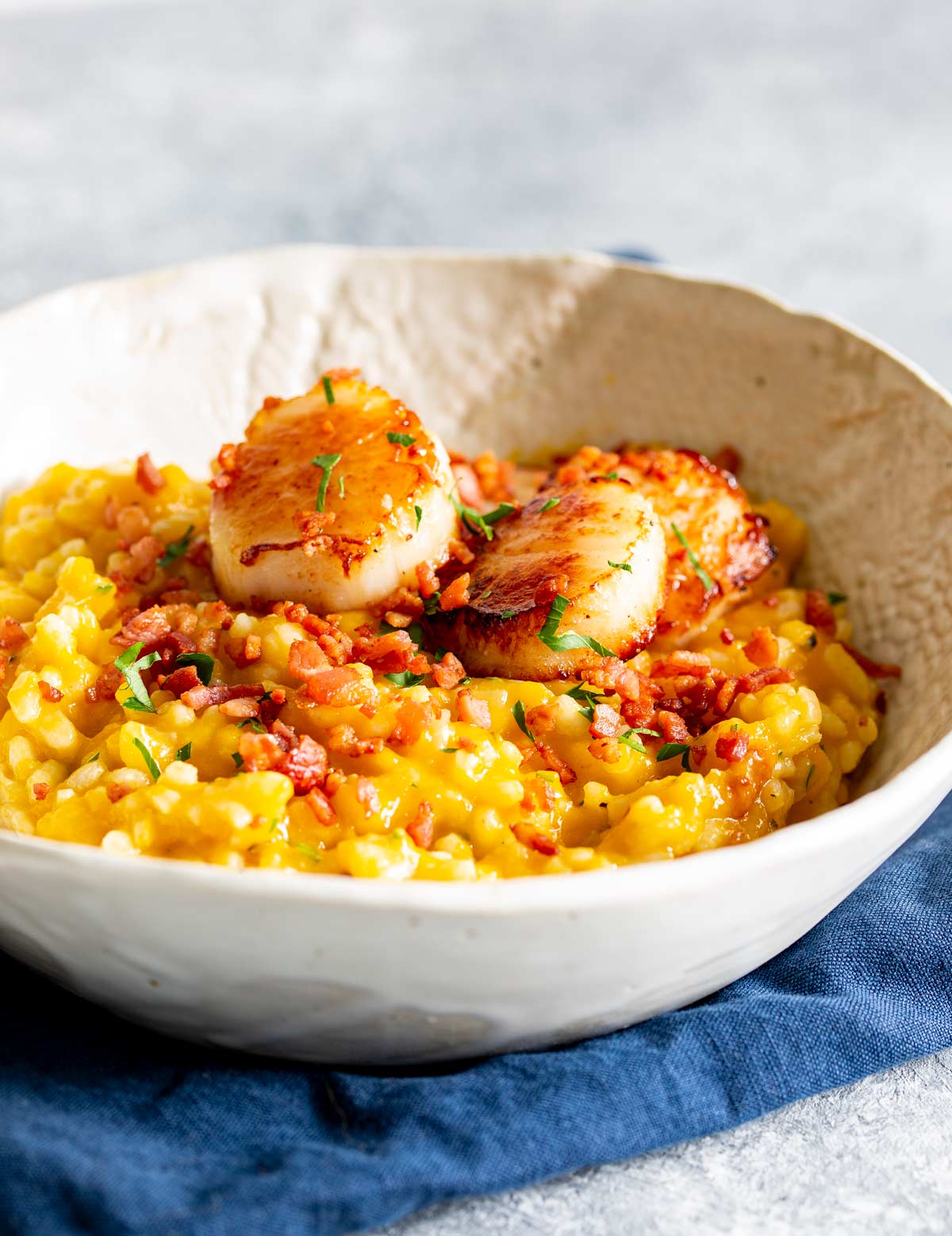 a rustic bowl filled with pumpkin risotto topped with bacon bits and seared scallops