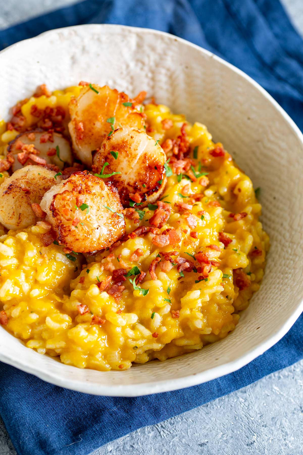 a scallop and pumpkin risotto in a cream bowl on a stone table with a blue napkin