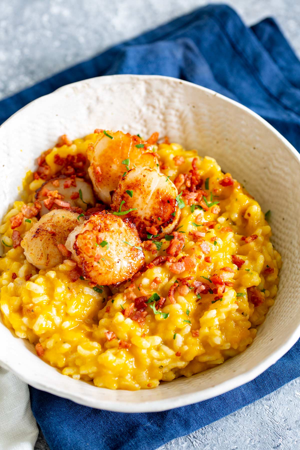 a cream bowl of risotto with bacon and scallops on top on a stone table with a blue napkin