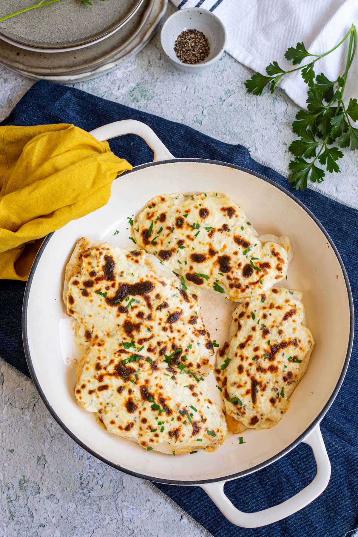 overhead of cheesy chicken breasts in a cast iron skillet on a table with a pile of plates