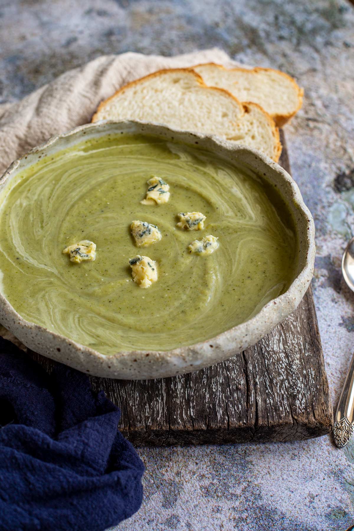 A rustic bowl of green soup on a wooden board on a stone table with bread behind