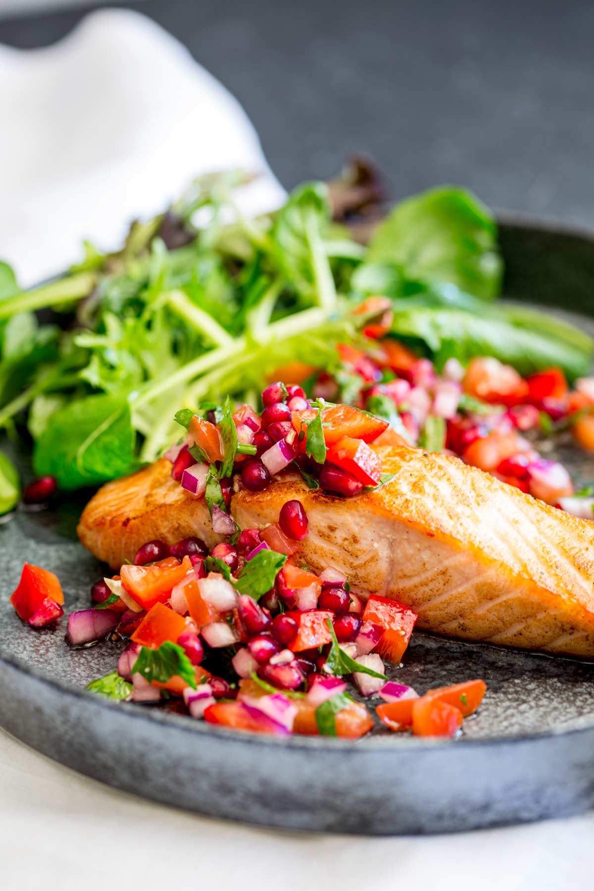 seared salmon with a pile of pomegranate salsa on it and green salad on a metal looking plate
