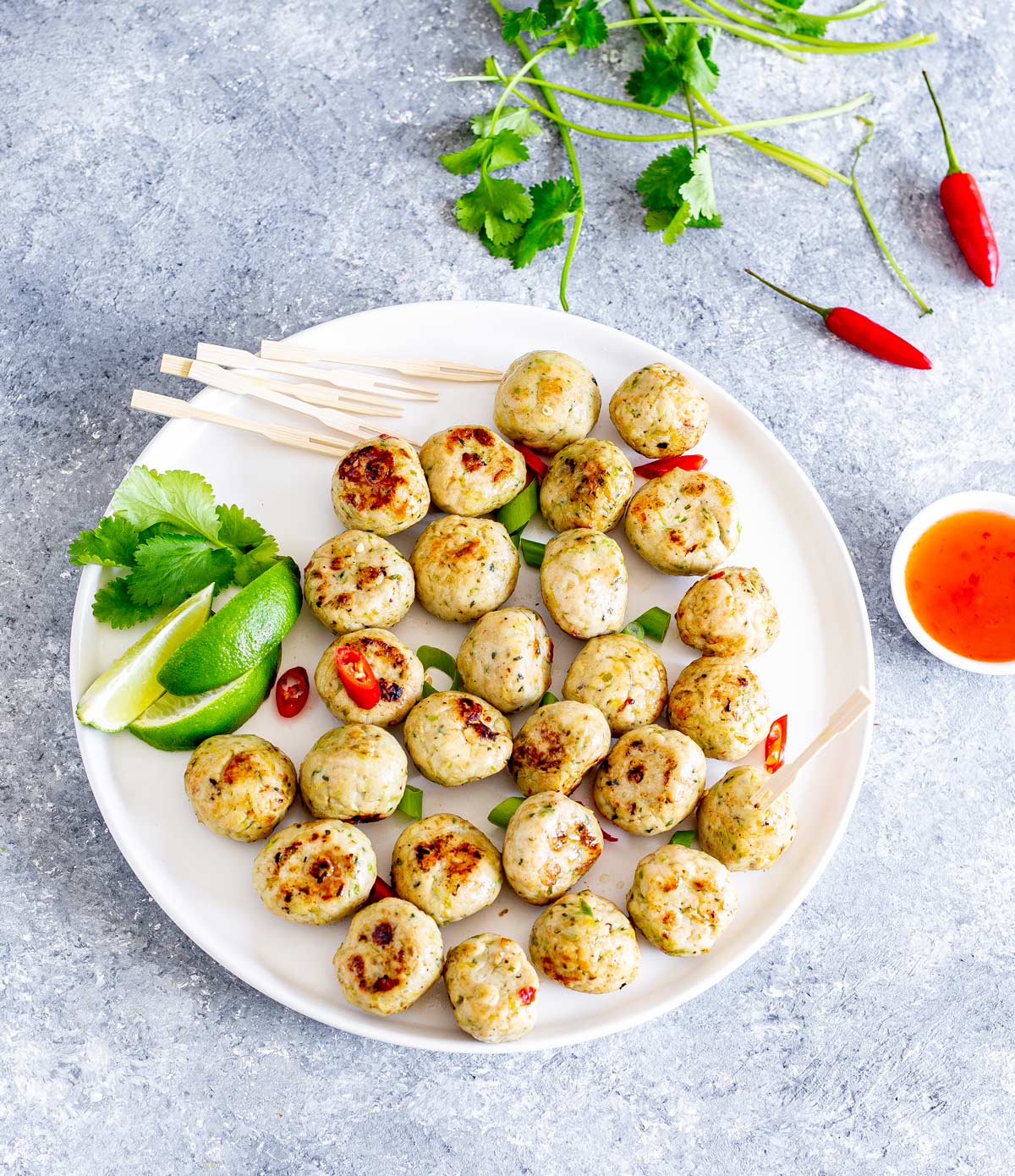 a white platter of Thai chicken meatballs garnished with sliced chili, green onions, lime wedges with a dish of chili sauce