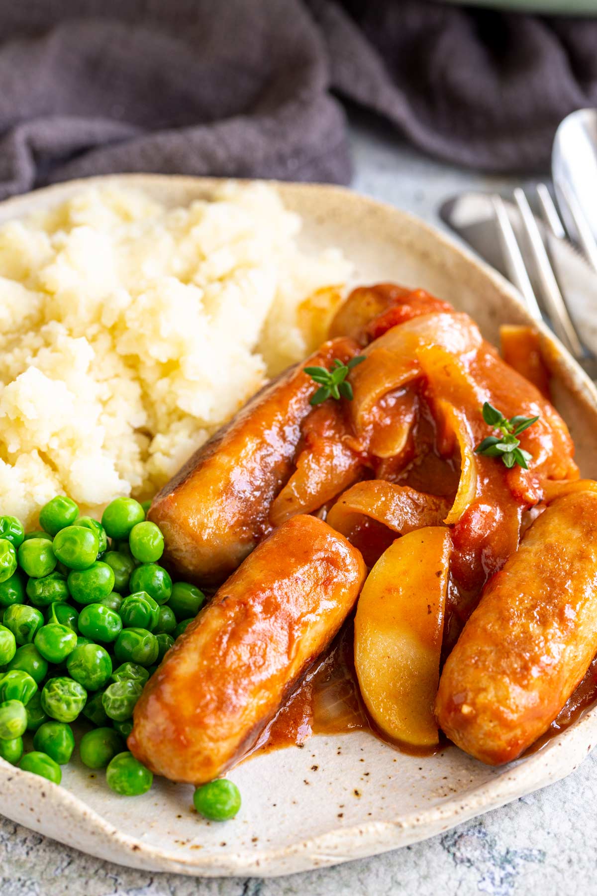 4 small sausages in a sauce on a plate with mashed potato and peas