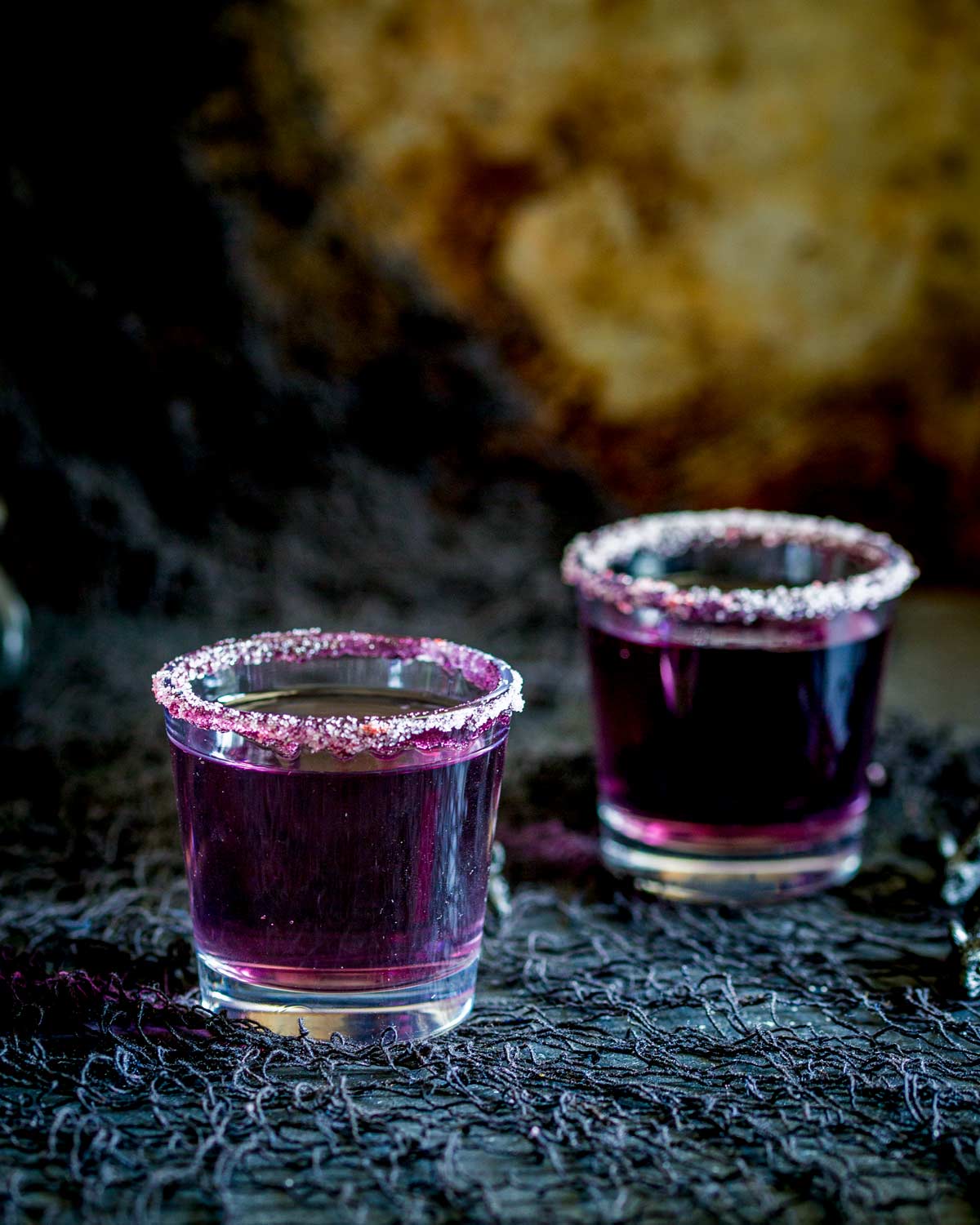 Two shot glasses with purple cocktails in them, on a black table.