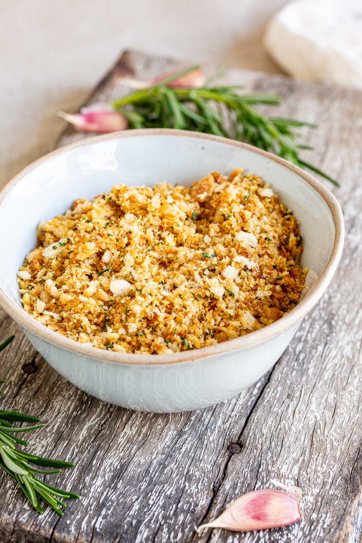 a rustic bowl of breadcrumbs on a wooden board with rosemary and garlic