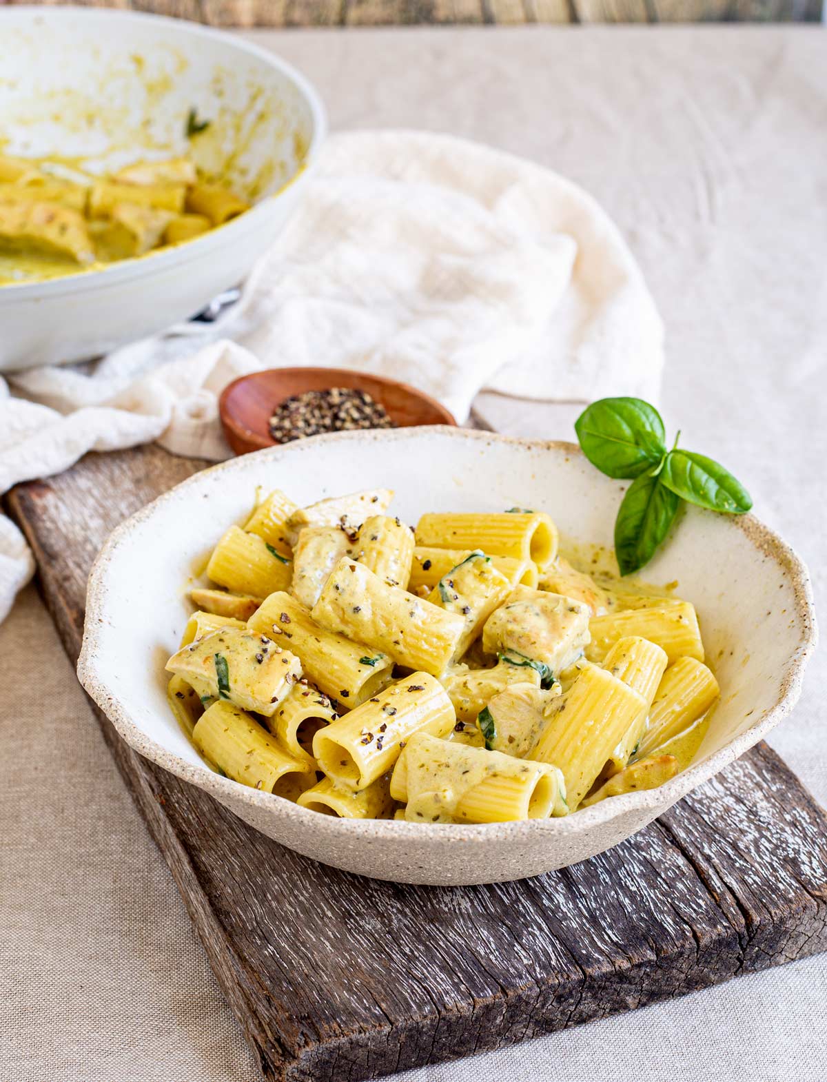 A rustic bowl of rigatoni pasta in a creamy pesto sauce on a rustic wooden board, garnished with fresh basil leaves. Behind is a skillet with the remaining pasta