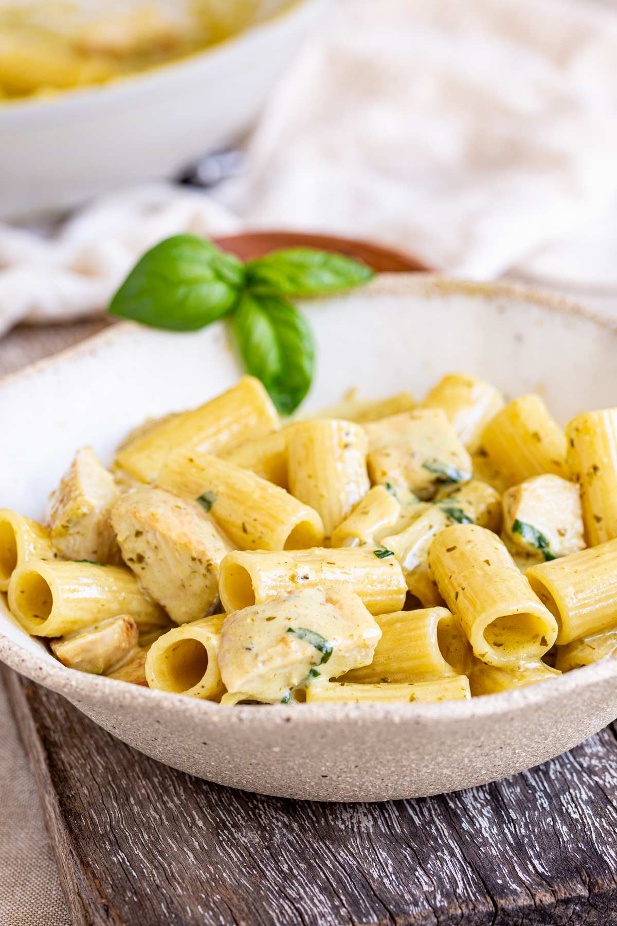 Side view of a white bowl, containing creamy pesto-coated pasta, chicken pieces, and a garnish of fresh basil leaves. The dish is styled with a soft focus background a linen napkin and another bowl of pasta