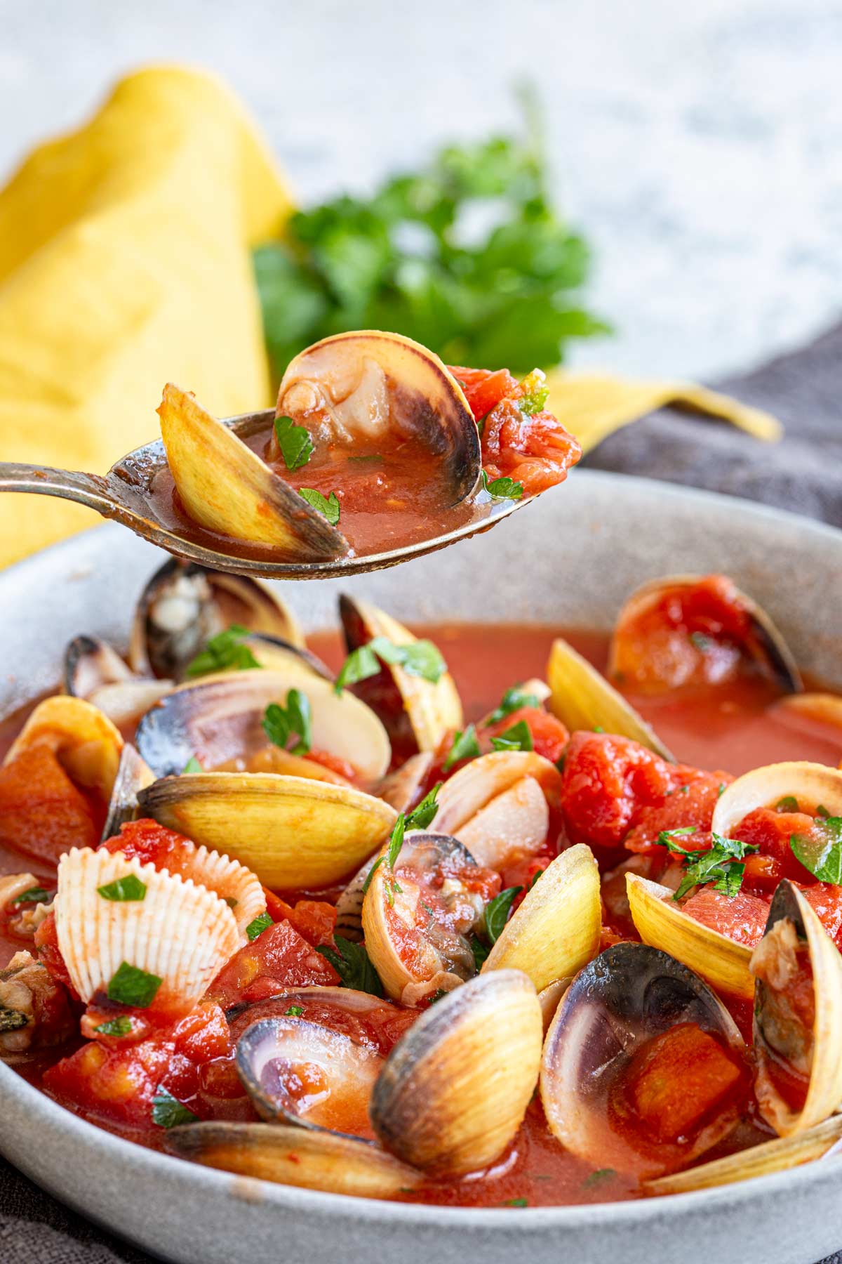 a spoon lifting up a clam and some tomato broth from a grey bowl