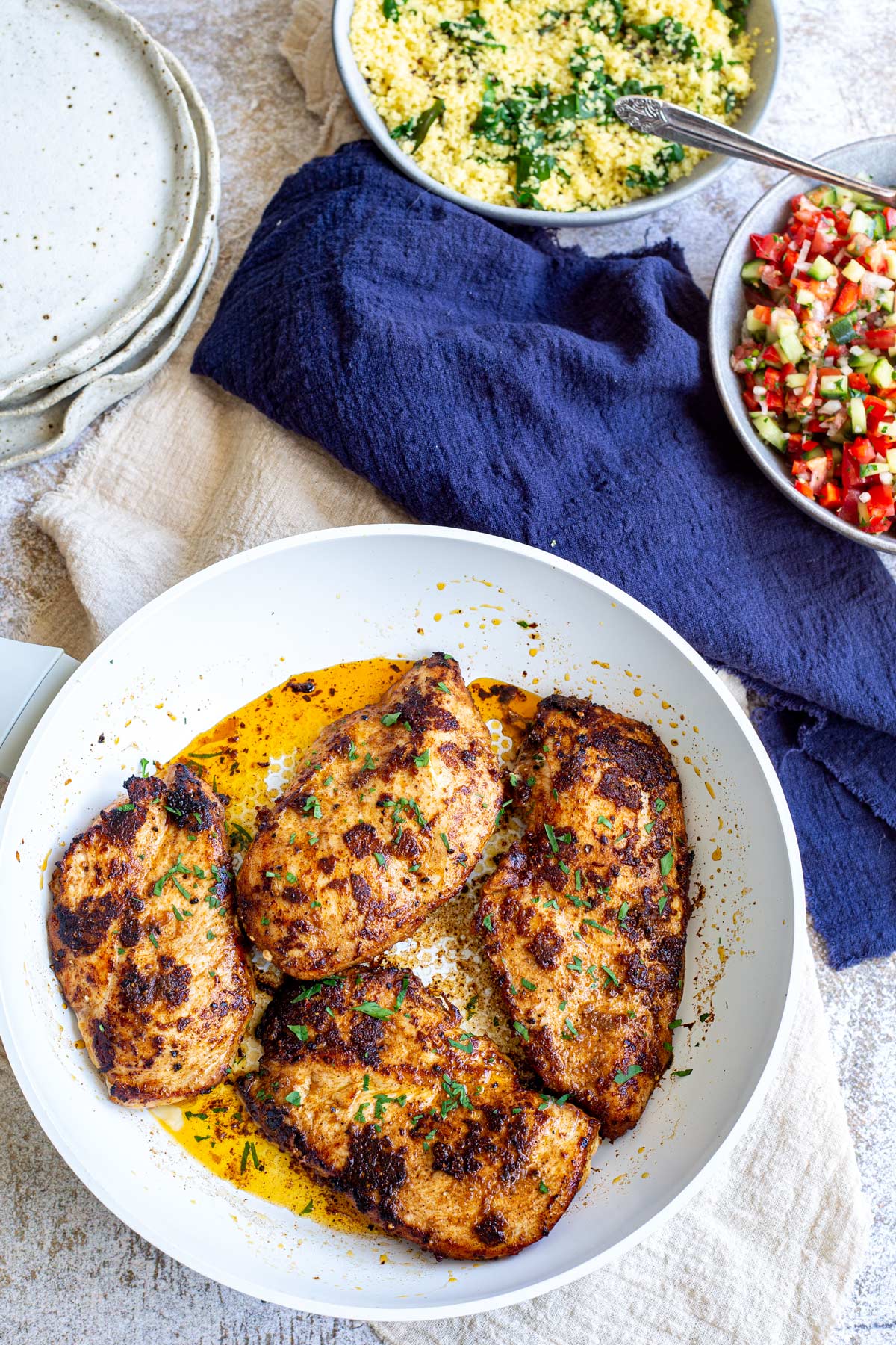 skillet with 4 chicken breasts, a bowl of tomato salad and a bowl of couscous on a table