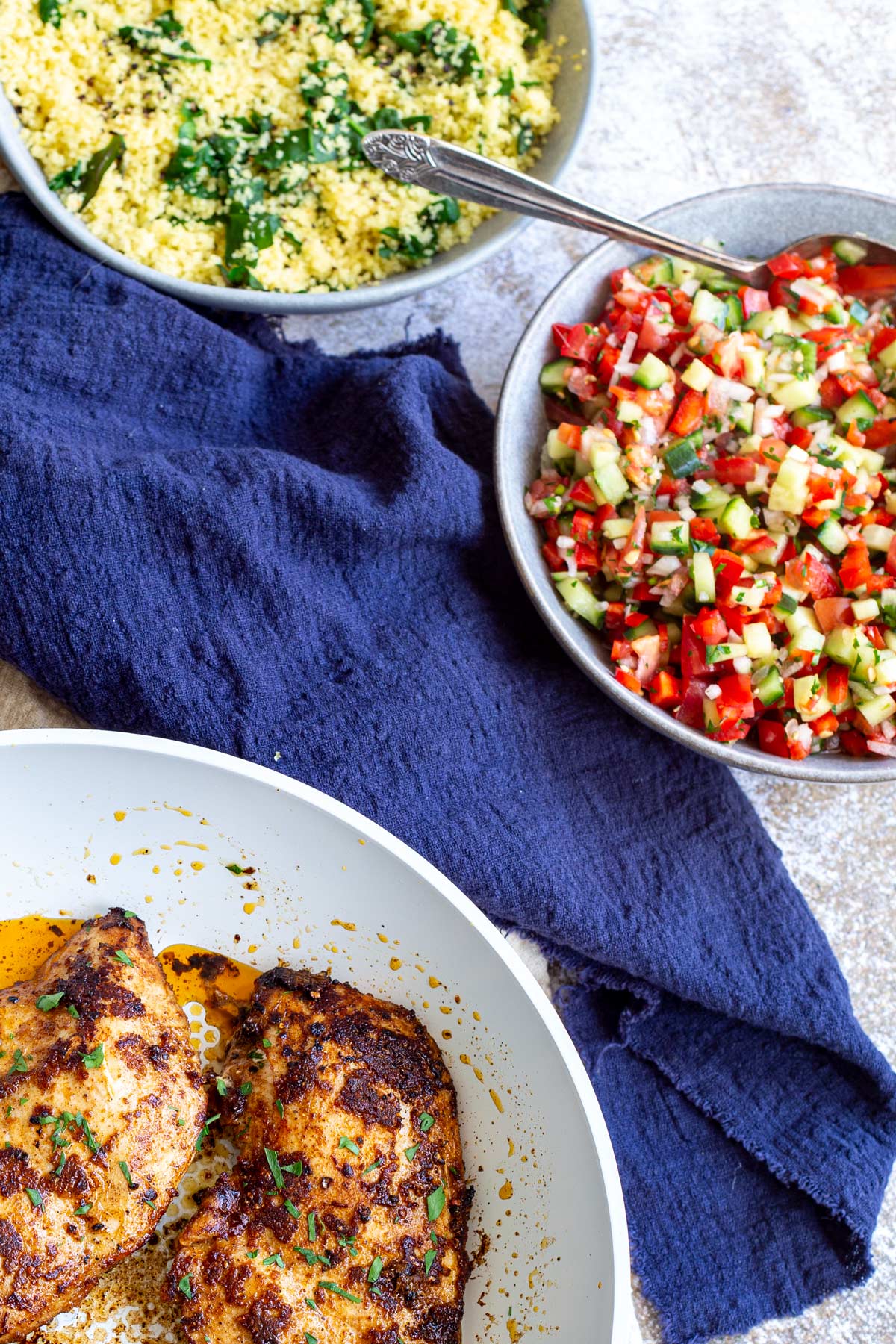 a sone table with a dish of tomato salad, couscous and a pan of cooked chicken breasts