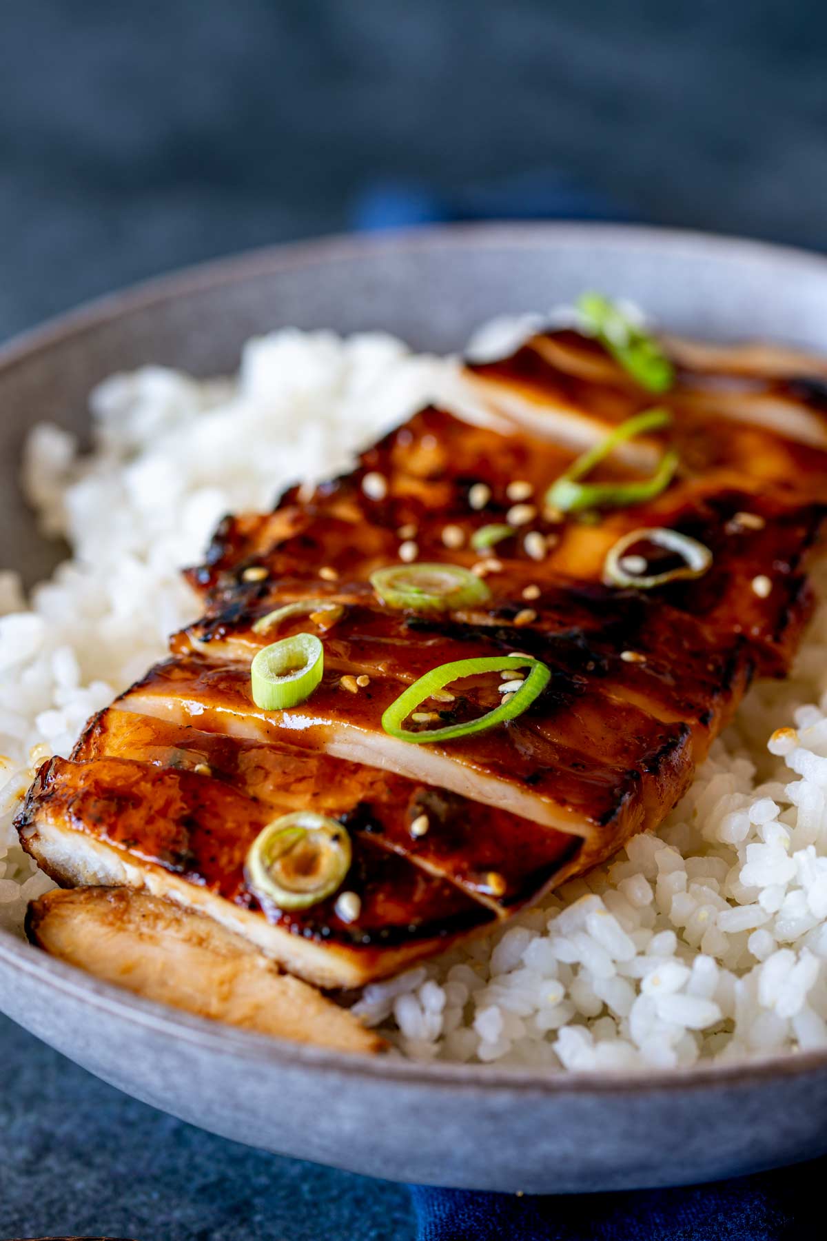 A sliced glazed chicken breast laid on sticky rice, topped with a sprinkling of sesame seeds and thinly sliced green onions