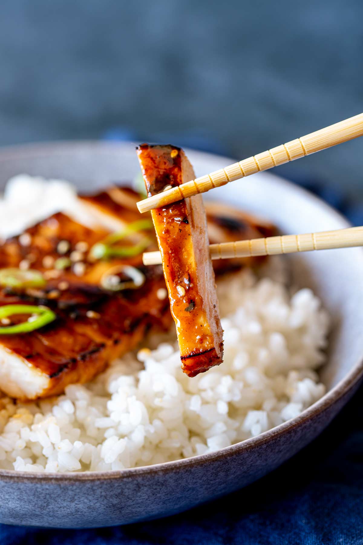 A close-up of a slice of chicken held by chopsticks, showing its glazed, caramelized surface.