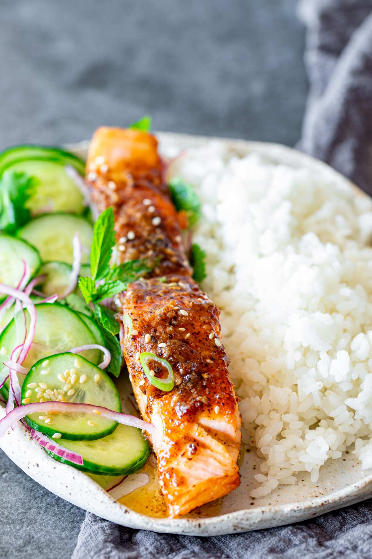 A serving of miso butter salmon fillet paired with sticky rice, cucumber salad, and mint leaves, arranged on a speckled ceramic plate.