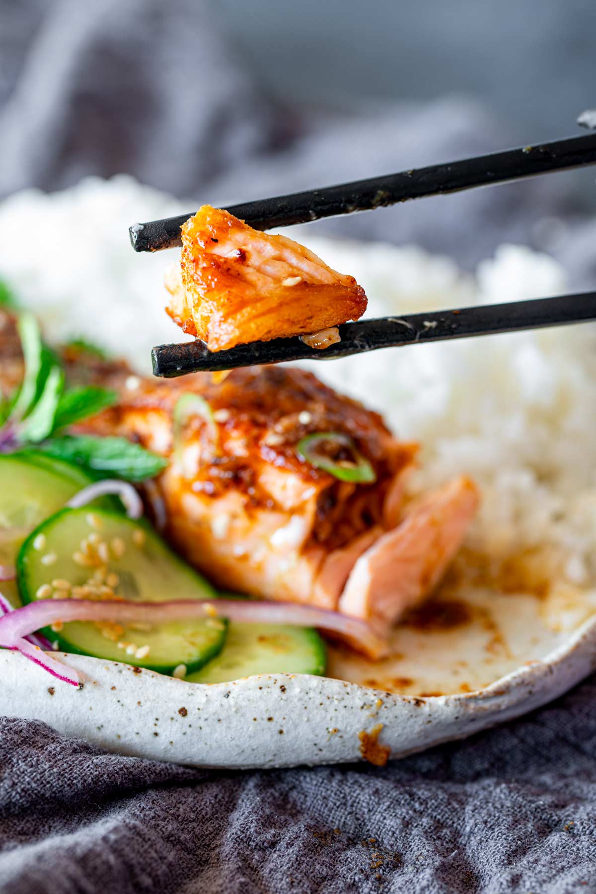 A piece of miso butter salmon held between black chopsticks with a side of rice and cucumber salad blurred in the background.