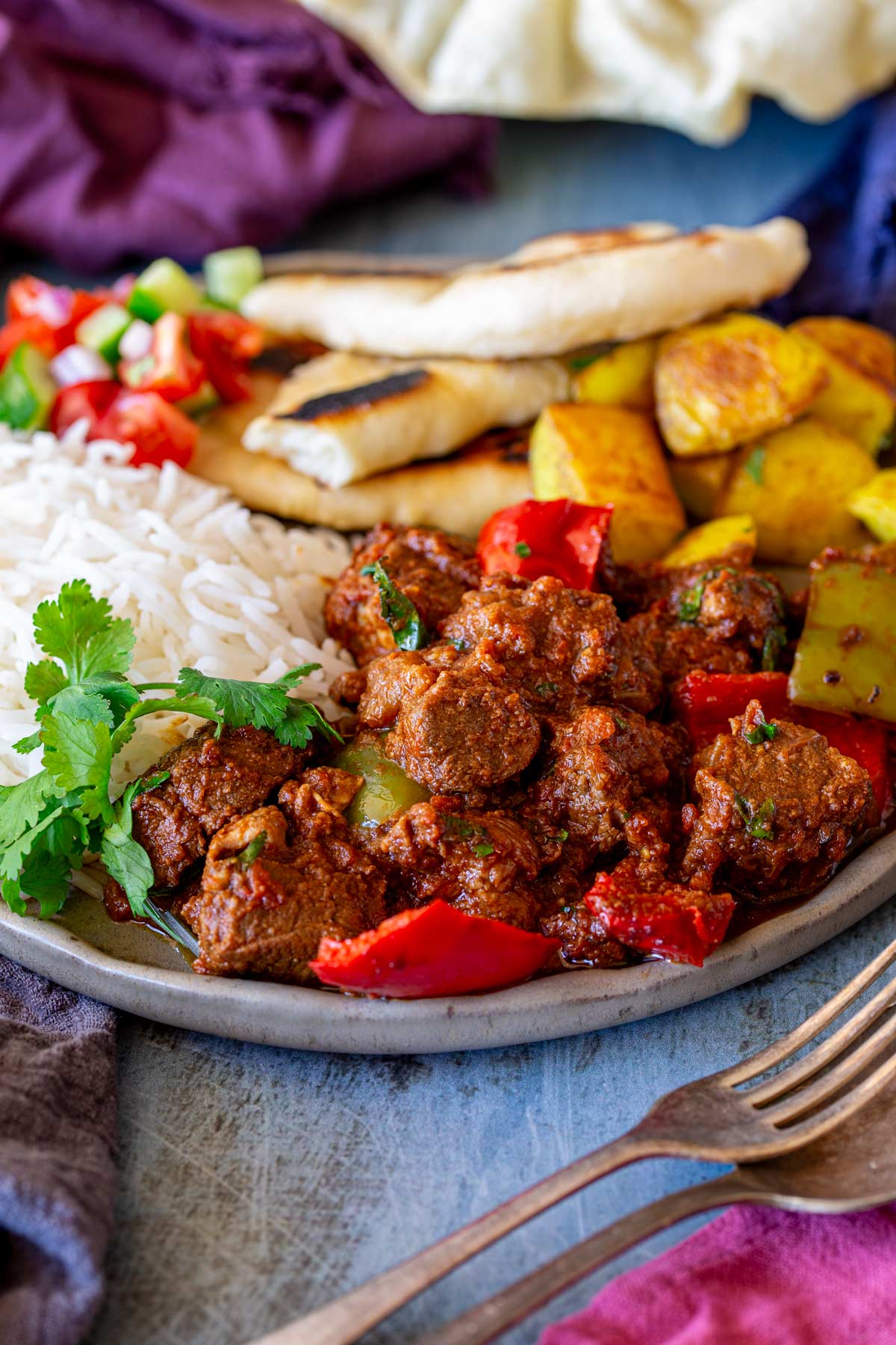 A plate of pork curry with red bell peppers, served with rice, Bombay potatoes, naan bread, and a kachumber salad.