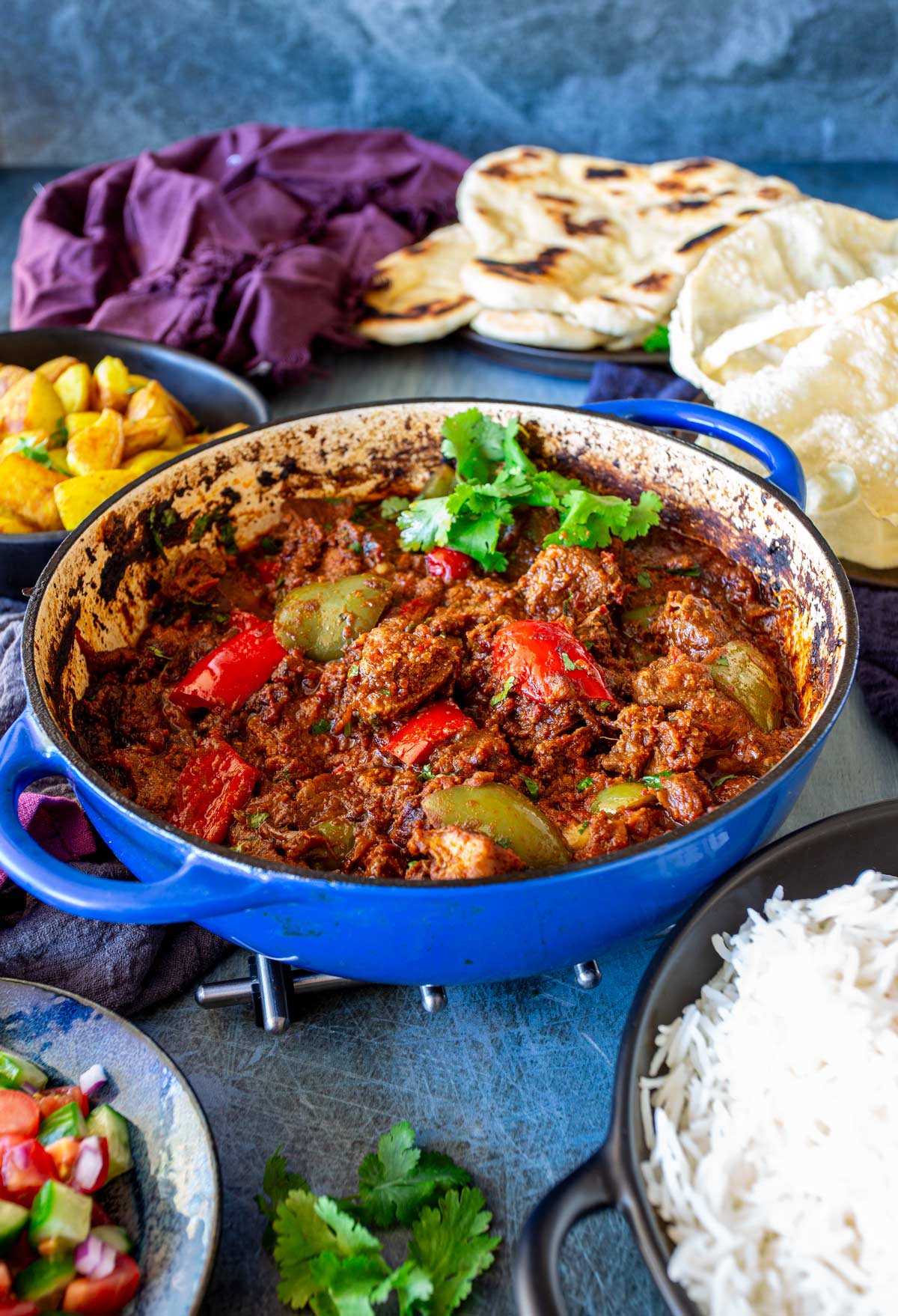 A blue pot filled with pork curry, surrounded by rice, naan, Bombay potatoes, and papadums