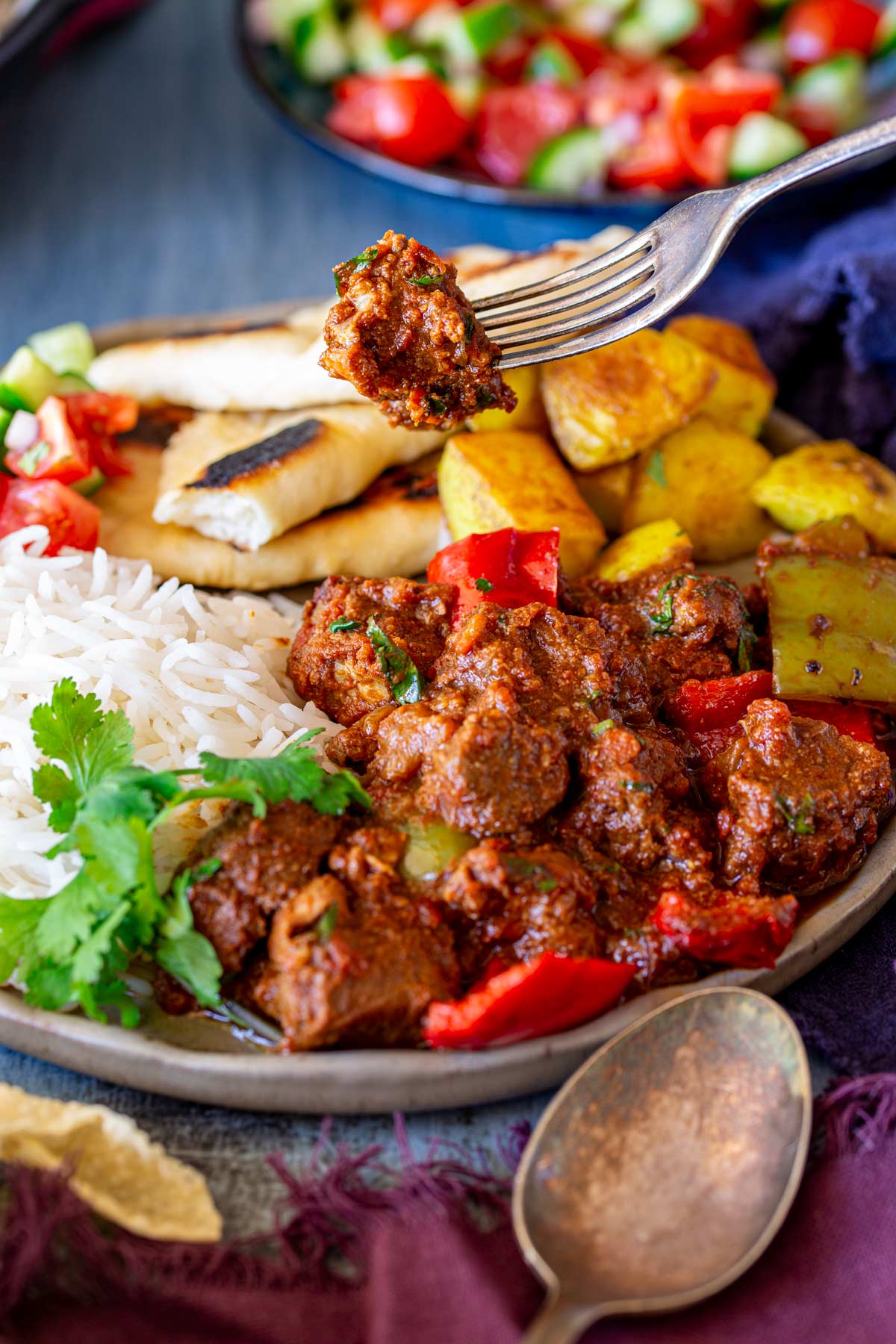 Fork lifting a piece of pork curry from a plate, featuring a colorful meal of curry, rice, naan, and Bombay potatoes