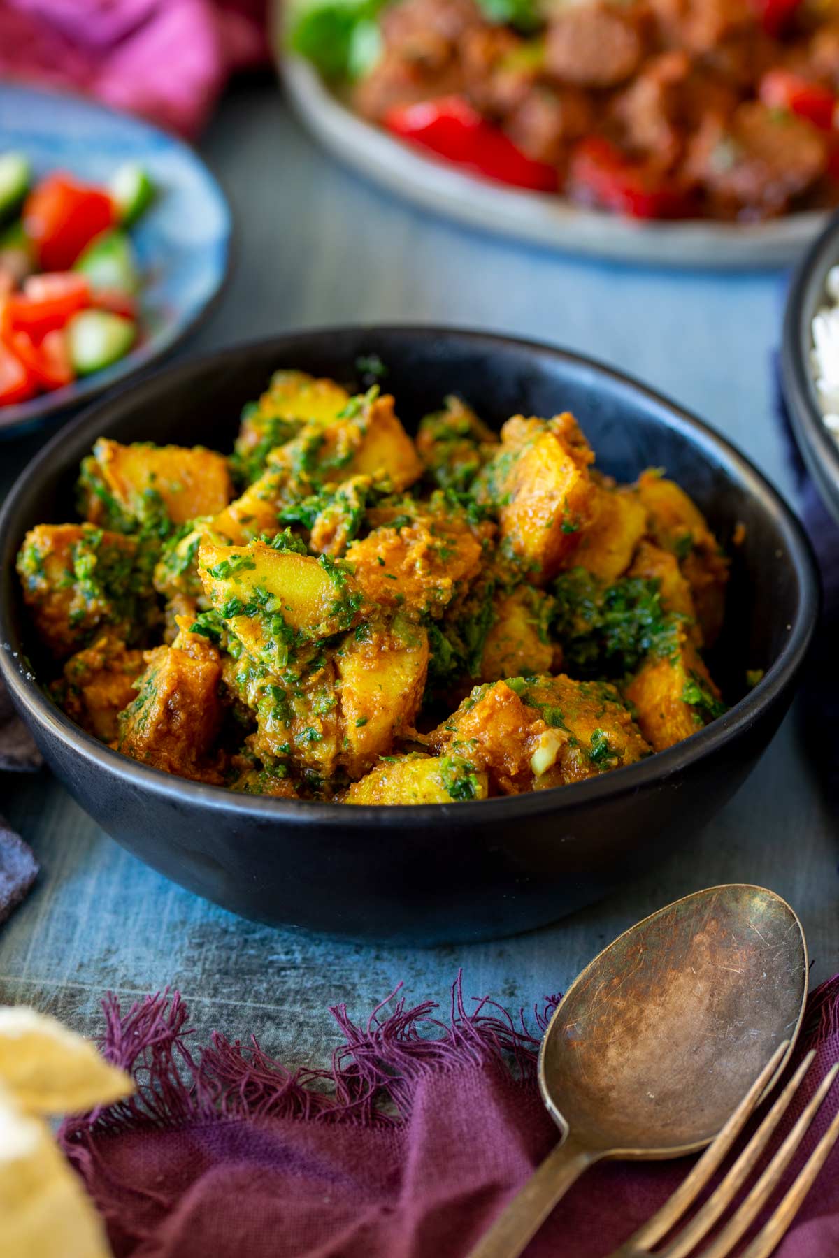 A black bowl filled with vibrant saag aloo, featuring golden potatoes and spinach in a spiced curry sauce, with a blurred background of salad and other dishes.