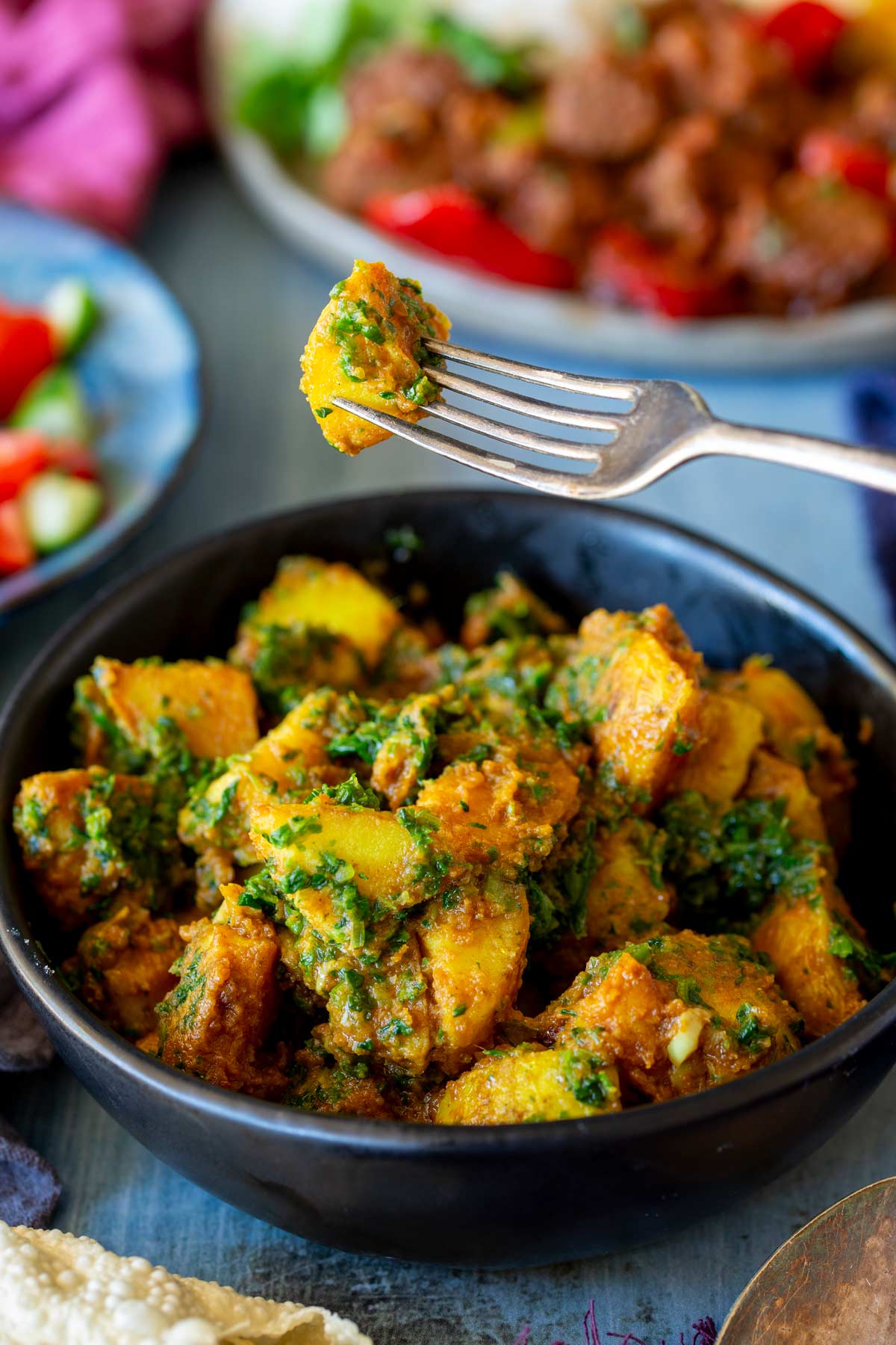 A fork lifting a piece of saag aloo, showing the texture of the potatoes and spinach, with more dishes and colorful salad in the background