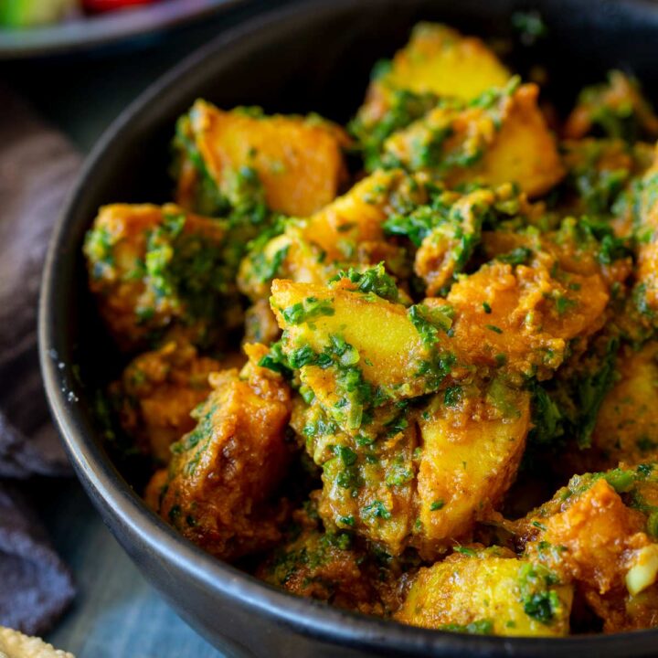 Close-up of saag aloo in a black bowl, with crispy papadum and salad visible in the background.