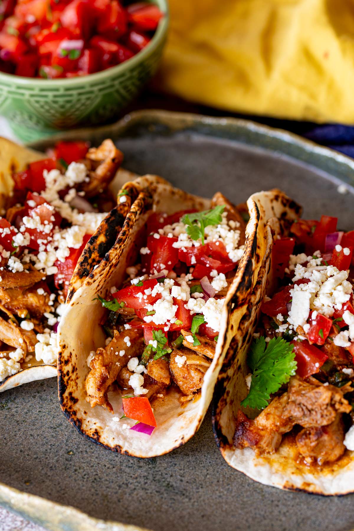 a dark plate with 3 chicken tacos on it garnished with queso fresco and tomato