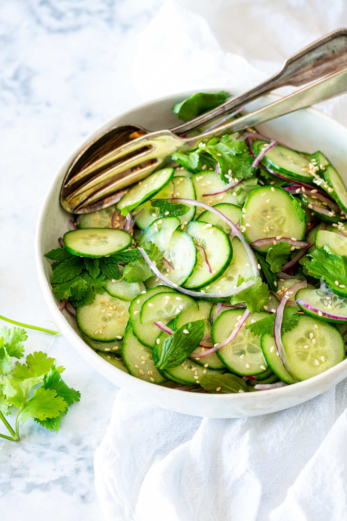 Fresh cucumber salad topped with red onion, cilantro, mint, and sesame seeds in a white bowl, with serving spoons resting inside.