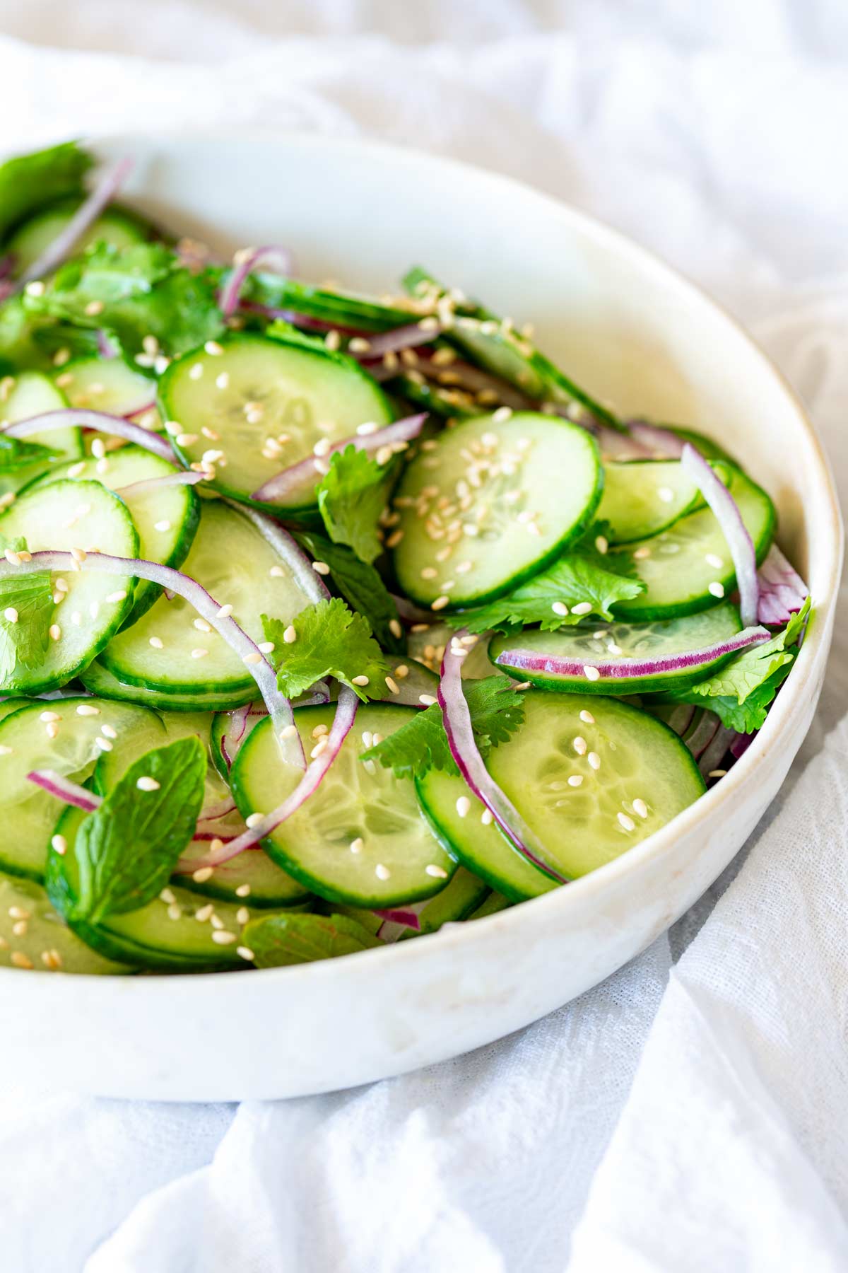 A bowl of cucumber salad garnished with fresh herbs and sesame seeds, and placed on a white cloth.