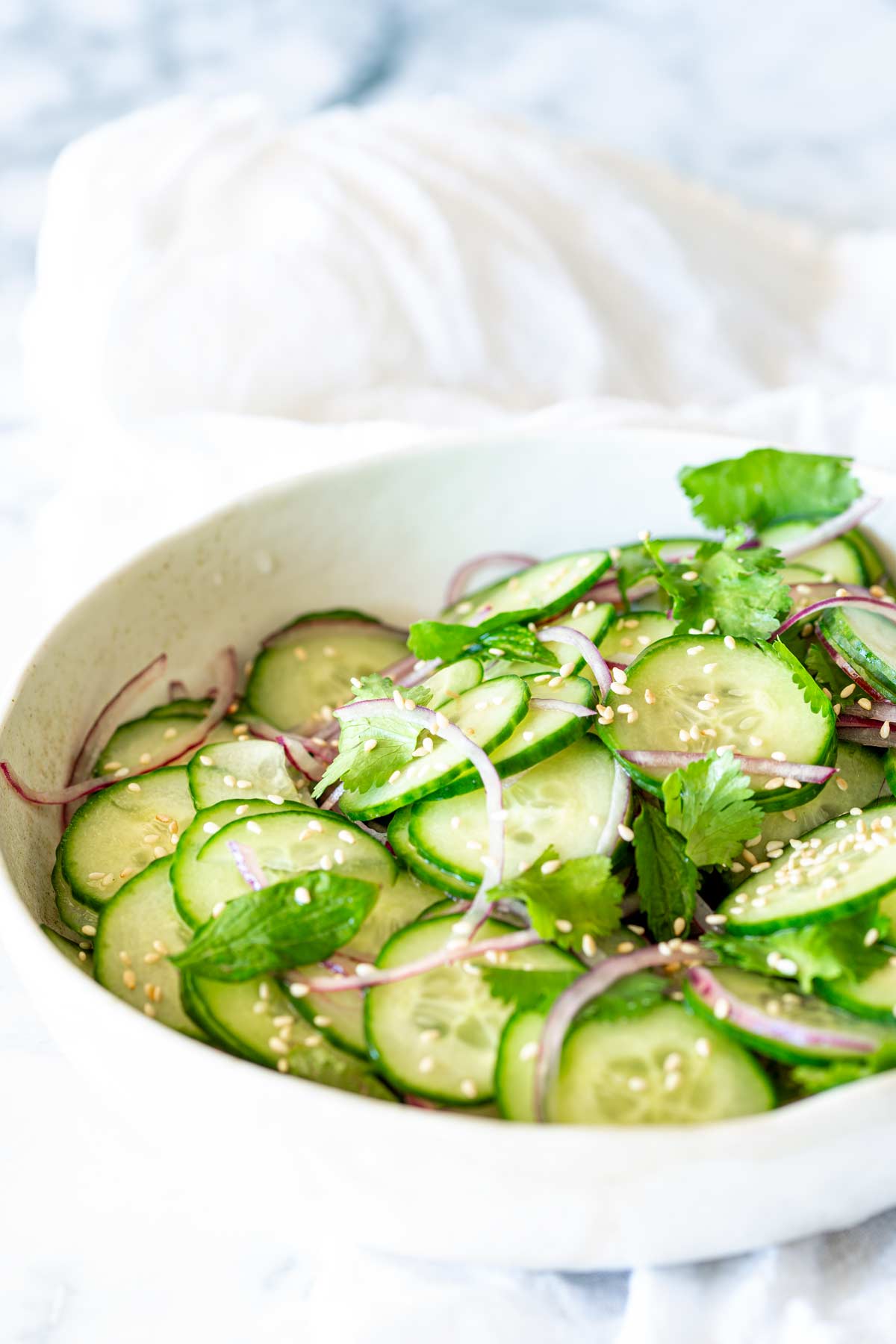 A cucumber salad with onions, cilantro, and sesame seeds, garnished with fresh herbs in a white ceramic bowl, placed on a white napkin.