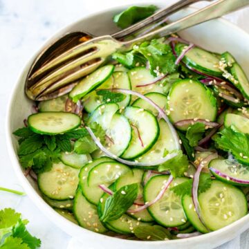 Fresh Asian cucumber salad in a white bowl, featuring thin cucumber slices, red onions, cilantro, mint, and sesame seeds, with a pair of serving spoons resting on the side.