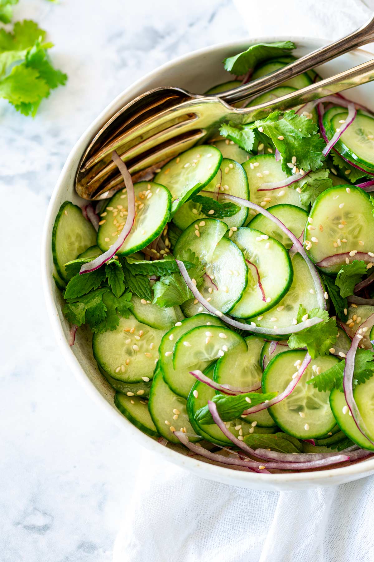 Close-up of an Asian cucumber salad with thinly sliced cucumbers, red onions, fresh herbs, and sesame seeds, served in a white bowl with serving utensils.