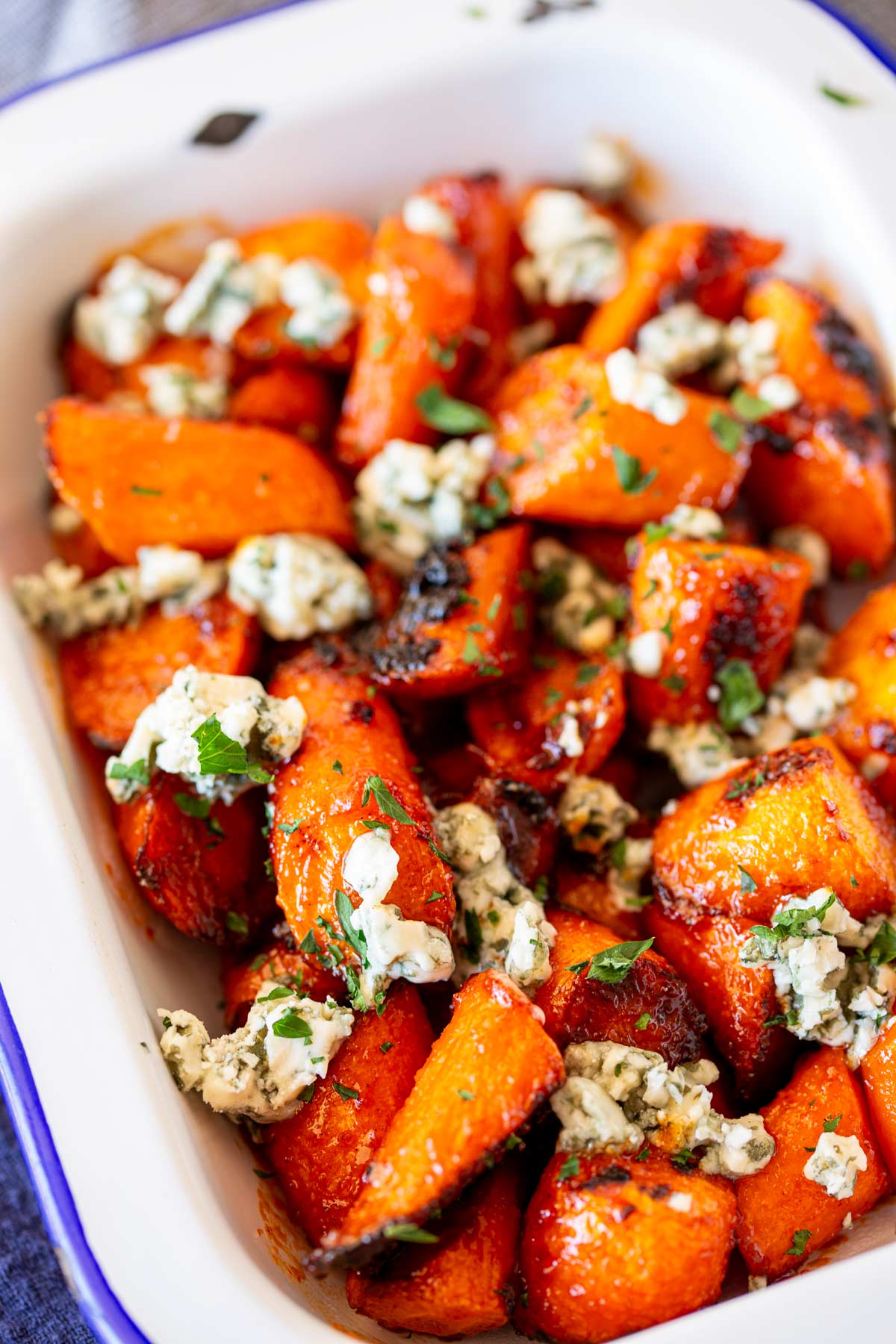 a rectangular baking dish filled with orange roasted carrots, coated in a rich buffalo sauce, and topped with white and blue crumbles of cheese, with green herbs sprinkled on top.