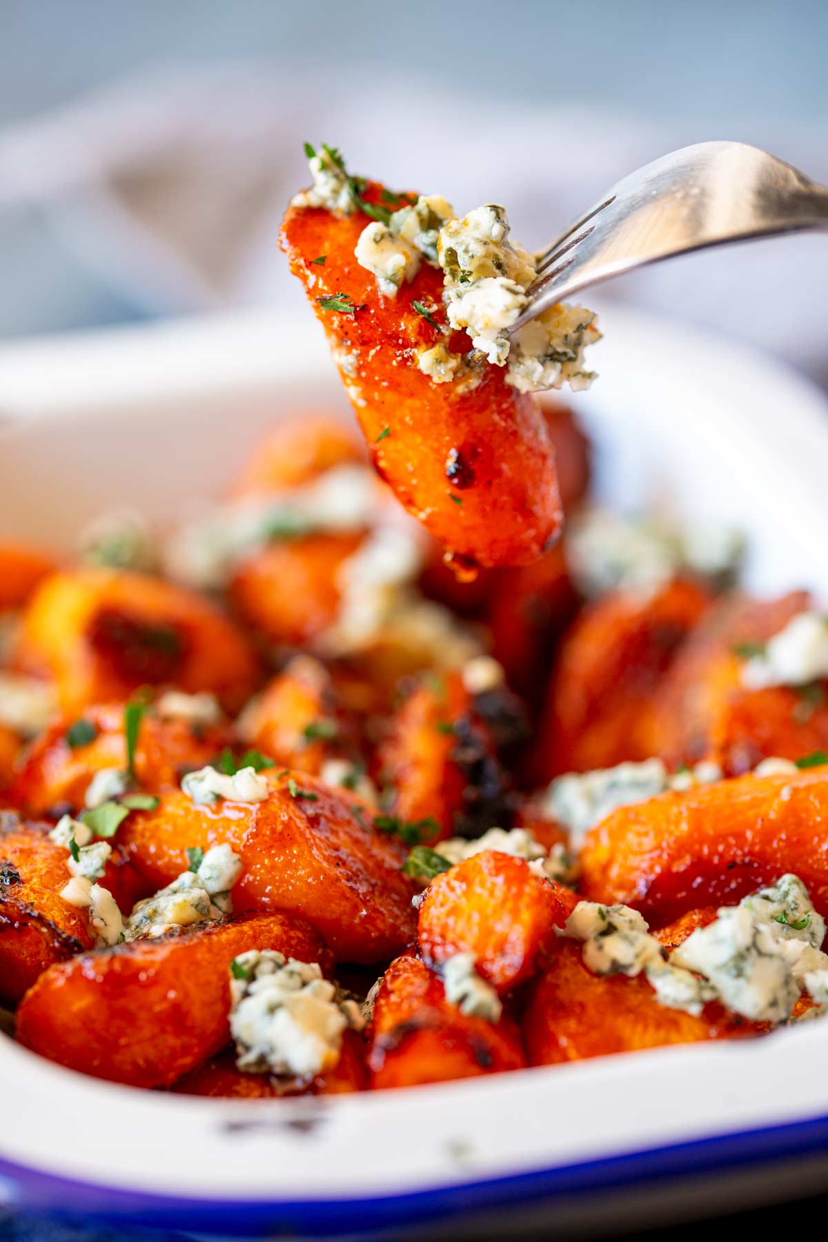 The image shows a fork lifting a roasted carrot wedge from a tray of buffalo-glazed carrots. The carrot is topped with a sprinkling of blue cheese crumbles, creating a mix of orange and white textures, and garnished with herbs.