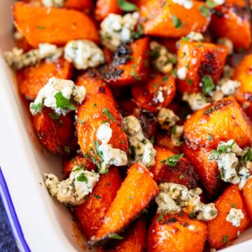 A baking dish filled with roasted carrots glazed in buffalo sauce, topped generously with crumbled blue cheese and garnished with fresh parsley.