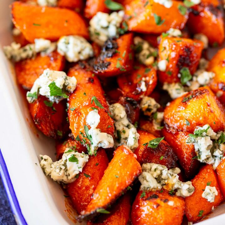 A baking dish filled with roasted carrots glazed in buffalo sauce, topped generously with crumbled blue cheese and garnished with fresh parsley.