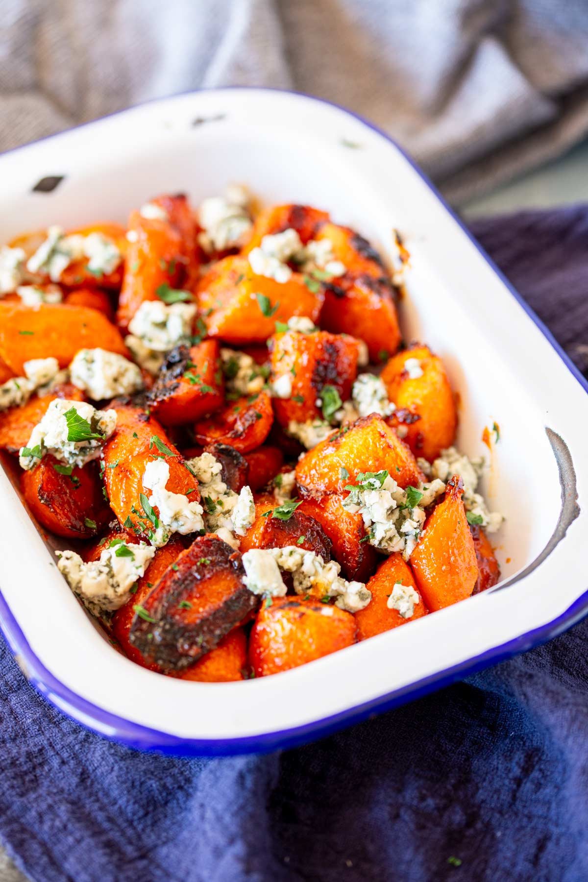 a white baking dish filled with roasted orange carrots, coated in a vibrant buffalo glaze. The carrots are topped with crumbles of blue cheese and sprinkled with herbs, all against a dark blue napkin