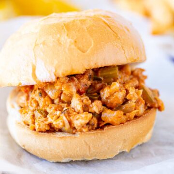 A toasted burger roll, with creamy, spiced chicken filling and bits of green pepper, sitting on parchment paper.