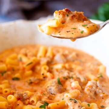 Spoon lifting creamy Italian sausage soup with pasta and sausage pieces, capturing a close view of the soup's ingredients, including parsley and tomatoes, in a speckled bowl.