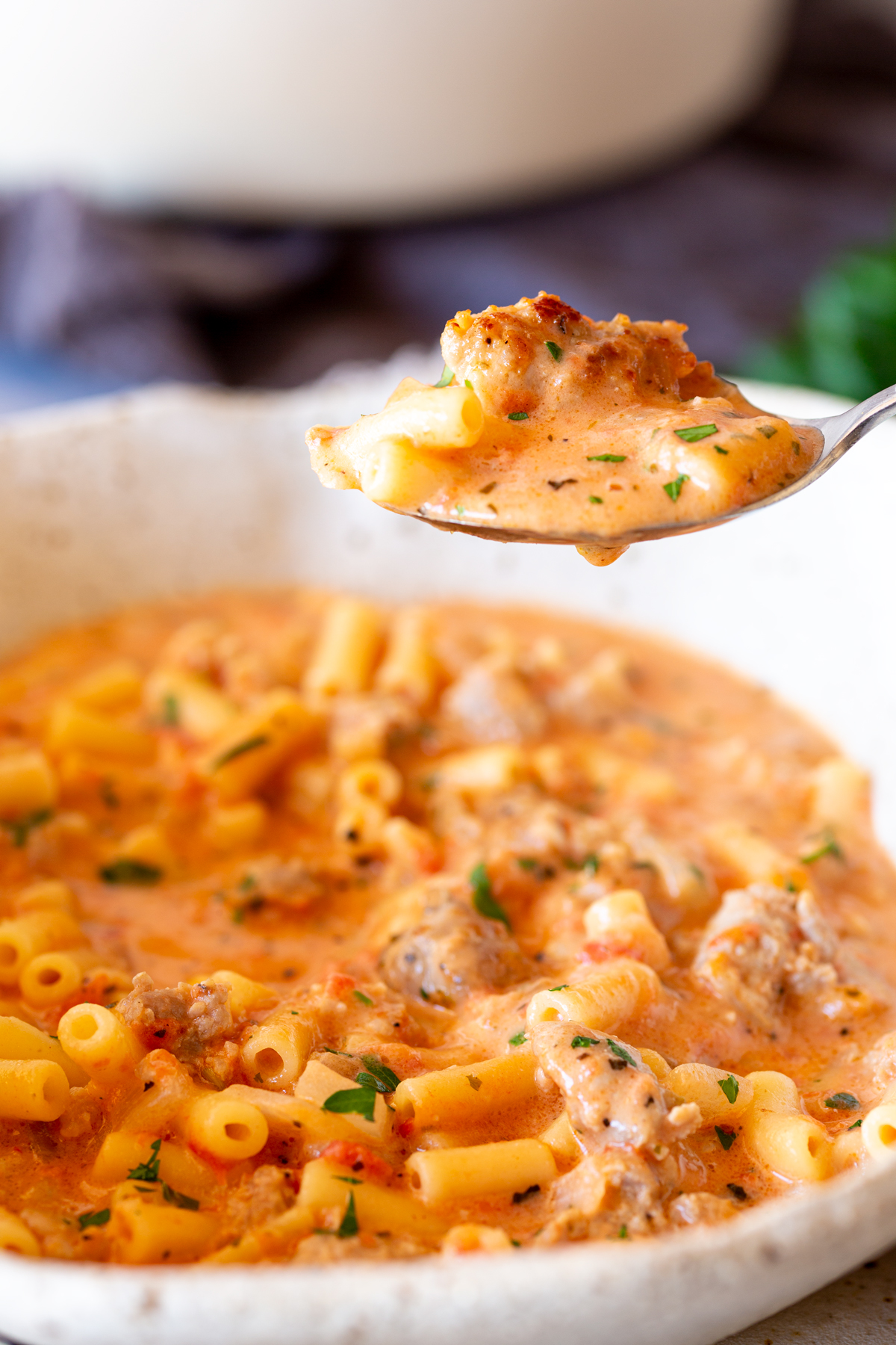 Spoon lifting creamy Italian sausage soup with pasta and sausage pieces, capturing a close view of the soup's ingredients, including parsley and tomatoes, in a speckled bowl.