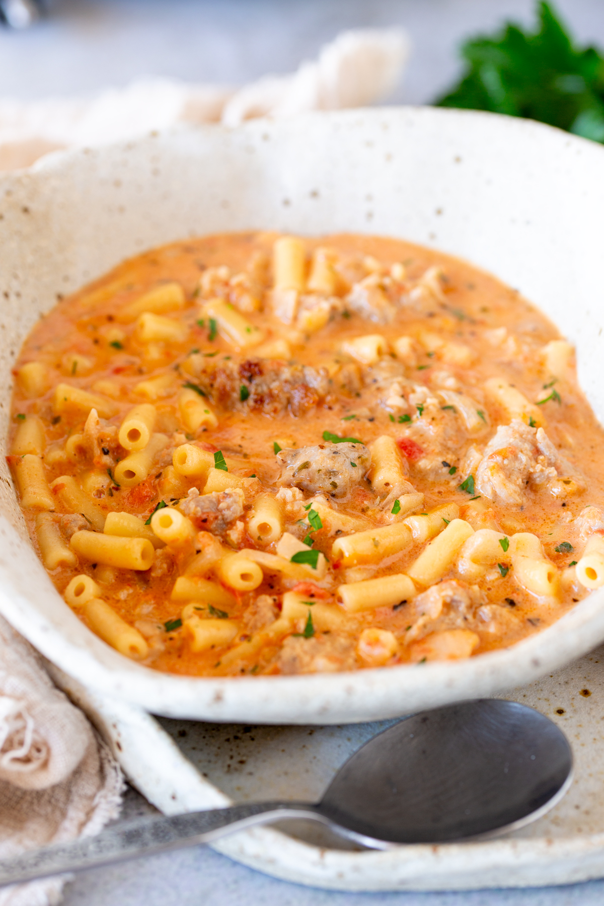 A bowl of creamy Italian sausage soup with pasta and sausage pieces, on a matching plate with a soup spoon
