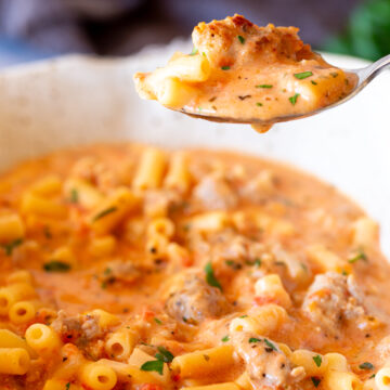 A close-up shot of a spoonful of creamy Italian sausage soup with small pasta tubes and chunks of sausage, showing a rich, hearty texture.