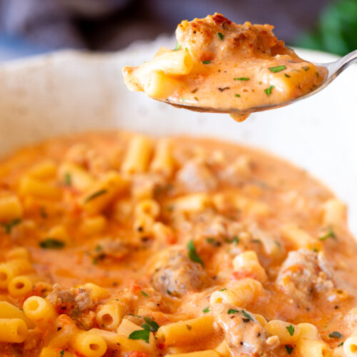 A close-up shot of a spoonful of creamy Italian sausage soup with small pasta tubes and chunks of sausage, showing a rich, hearty texture.