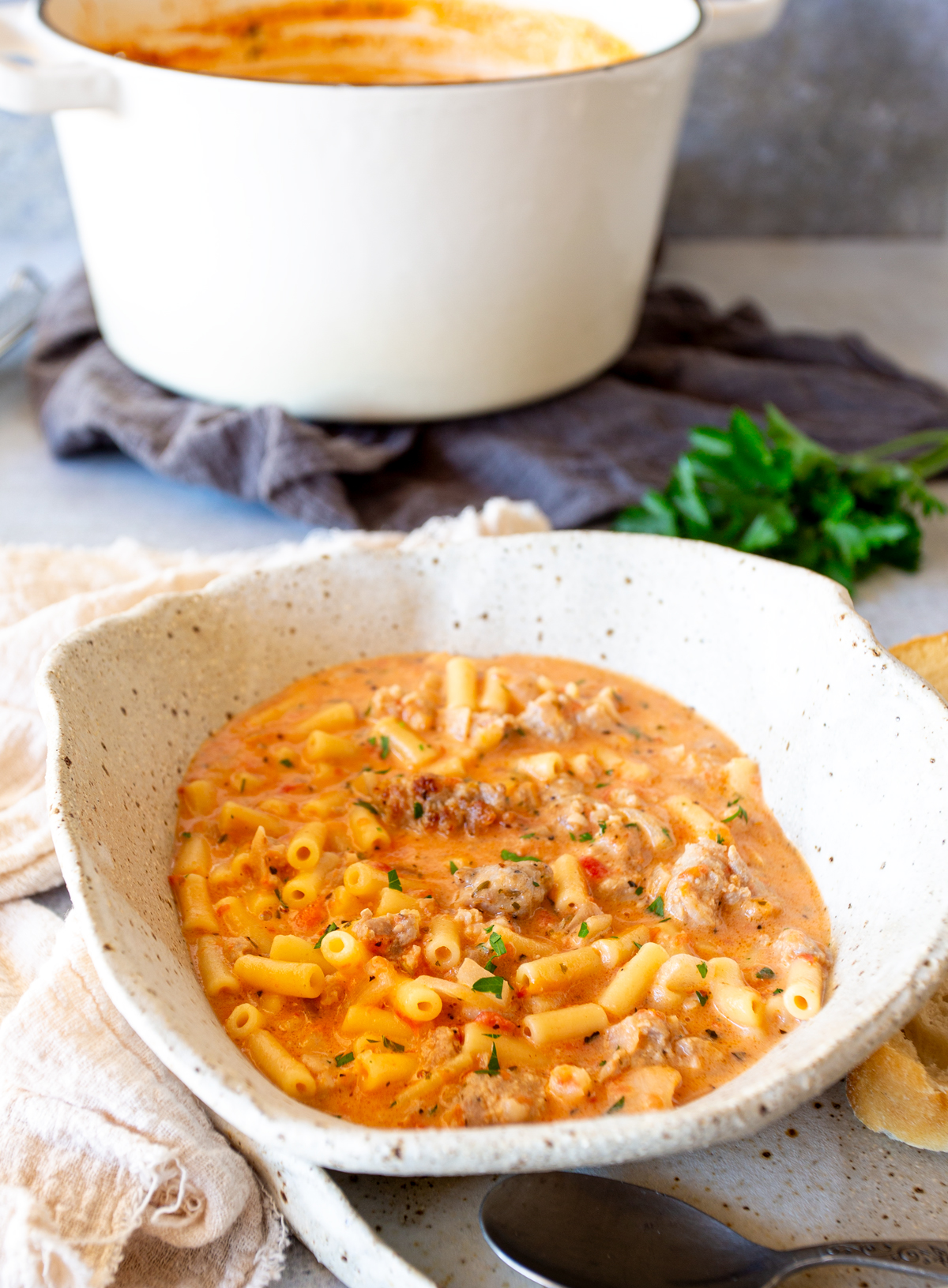 Speckled ceramic bowl filled with creamy Italian sausage soup with small pasta tubes and sausage chunks, served with fresh parsley and placed on a cloth with the cooking pot nearby.