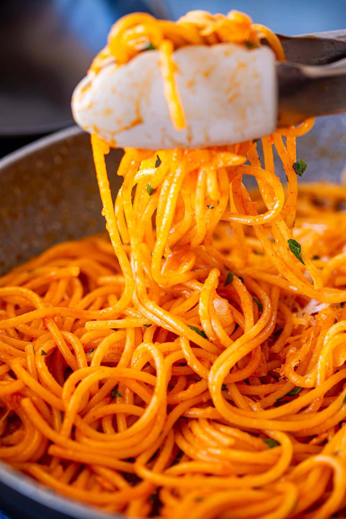 A pair of tongs lifting spaghetti from a pan, with each strand evenly coated in an orange-red gochujang butter sauce. Small green herb bits add color contrast.