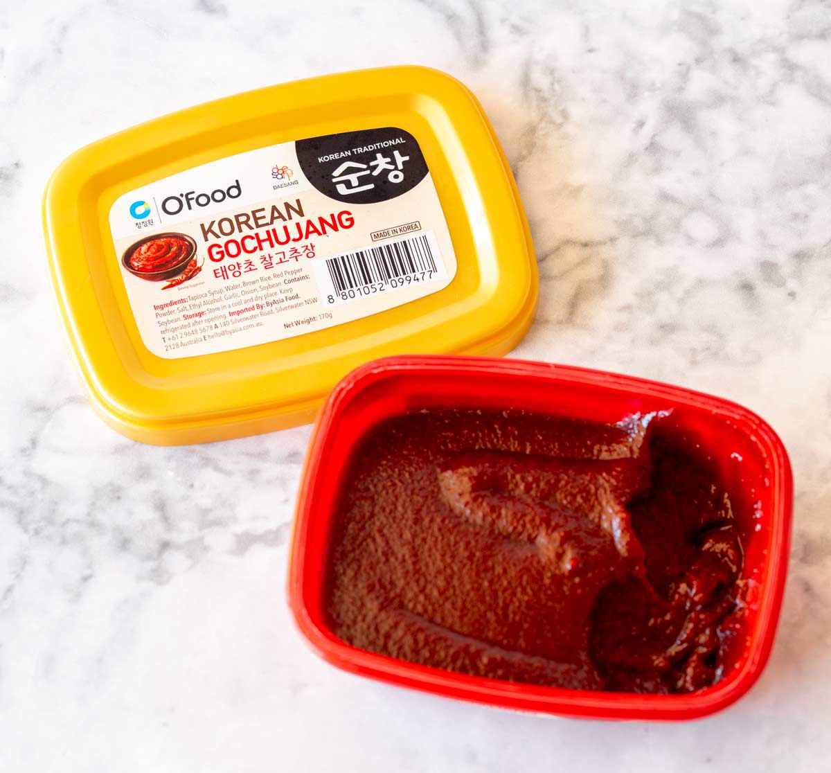 a red tub of gochujang paste with the yellow lid taken off and placed beside the tub on a marble countertop