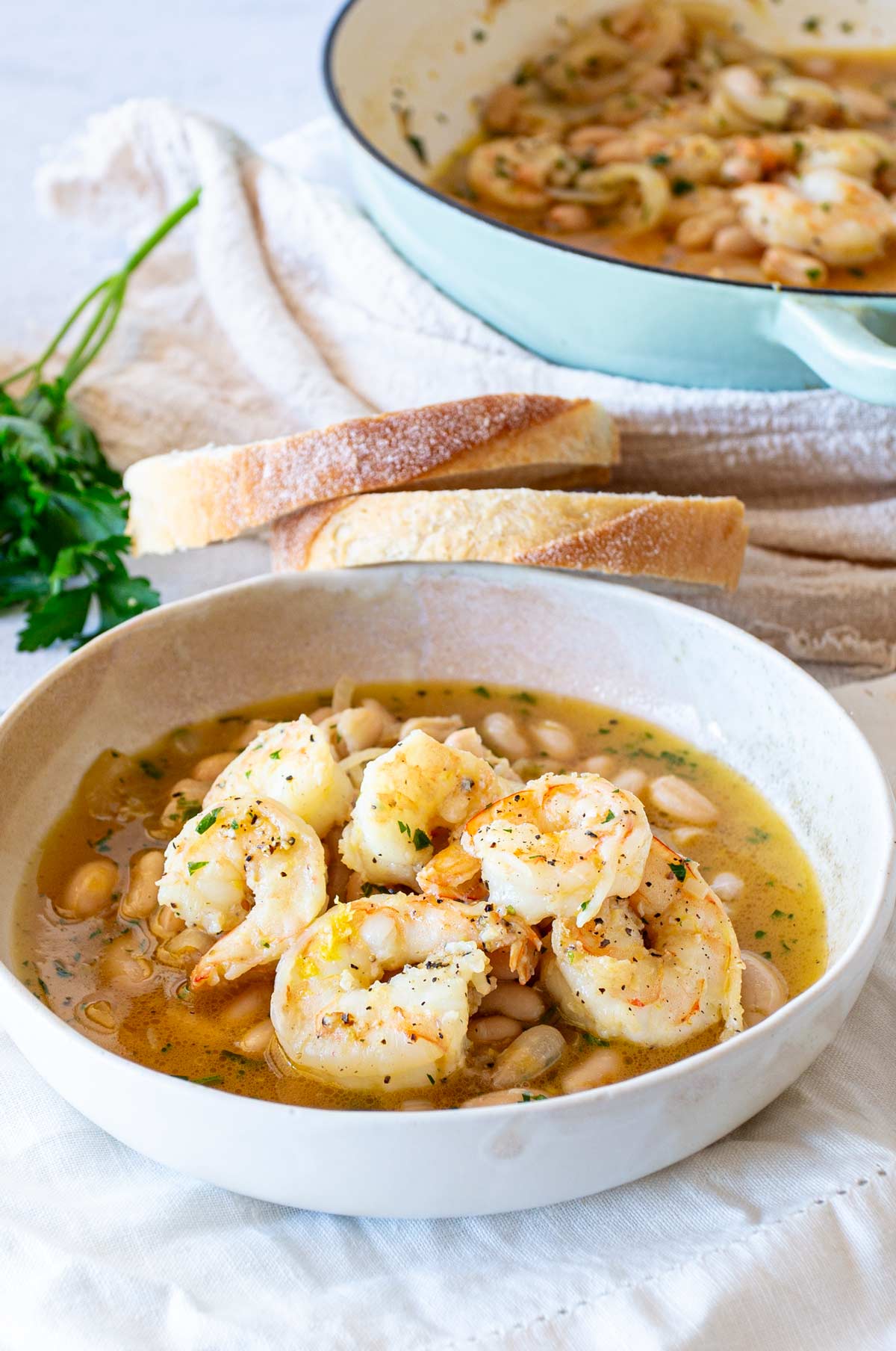 A beige ceramic bowl containing juicy shrimp, cannellini beans, chopped herbs, and a light lemony broth. Behind the bowl, there's a green Dutch oven and slices of baguette resting on a linen cloth.