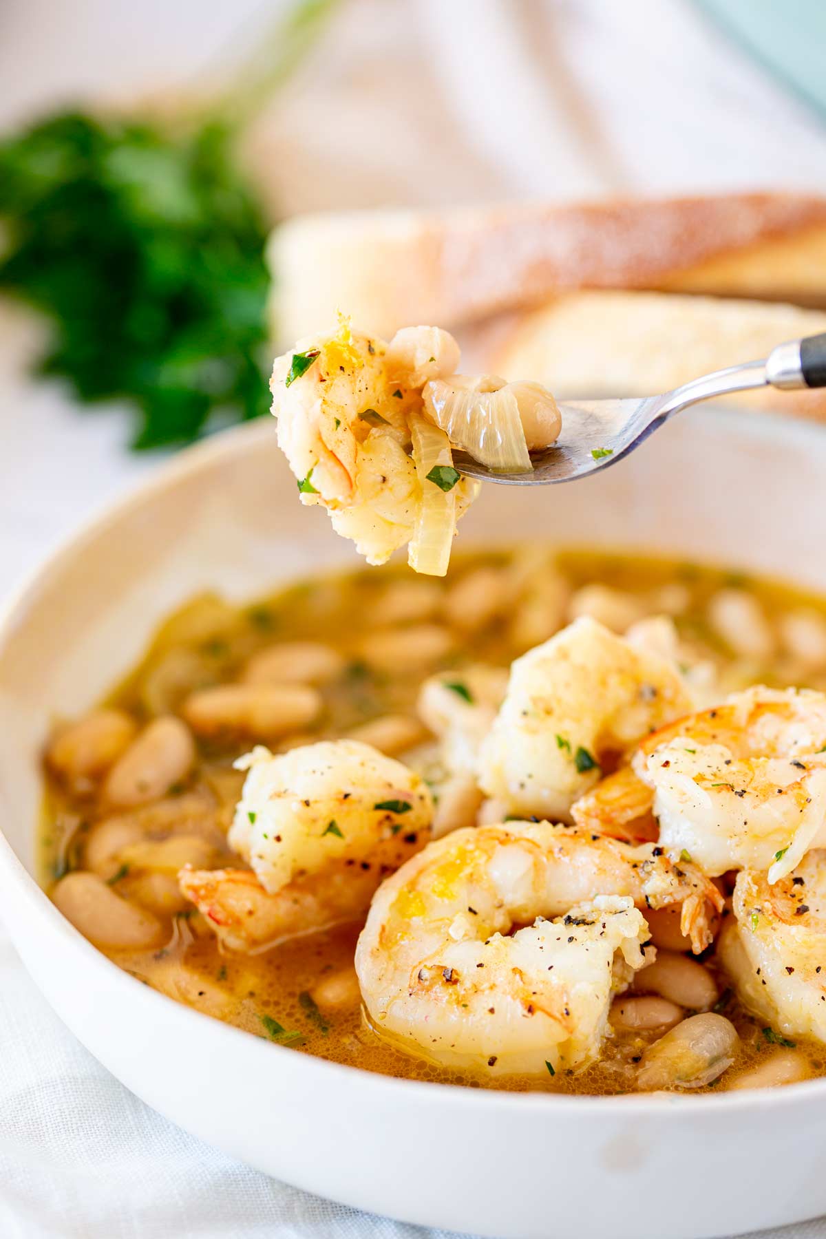 A fork holding a plump shrimp with sautéed onions, fresh parsley, and creamy white beans in a light broth, with parsley and crusty bread blurred in the background.