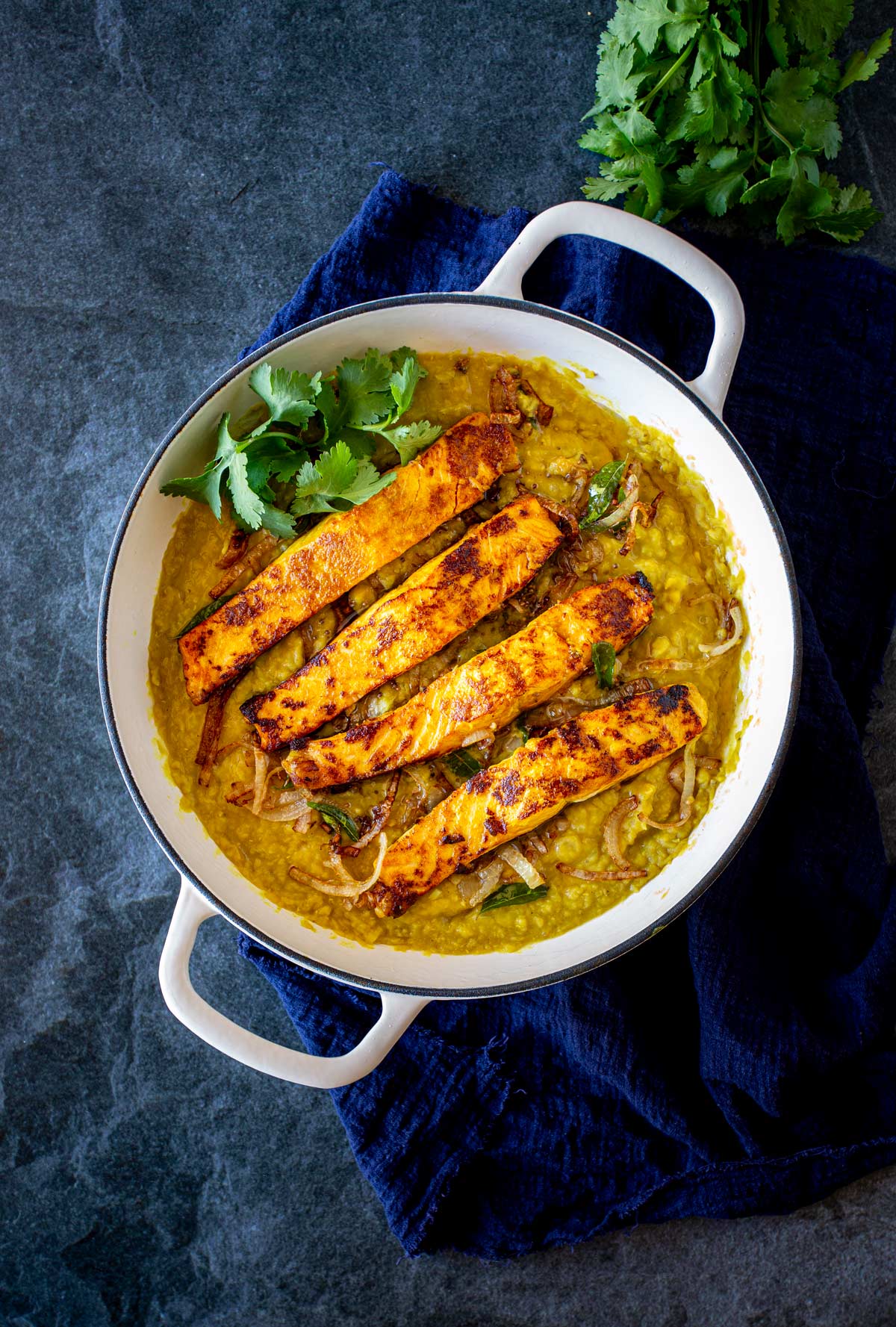 A white skillet filled with golden lentil daal, topped with seared salmon fillets and garnished with fresh cilantro, set on a dark surface with a navy blue cloth underneath.